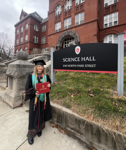 Graduate stands next to Science Hall wearing graduation robe and cap with yellow cords and a blue ribbon, holding red diploma and smiling.
